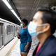 Two people wearing surgical masks wait at a subway station.